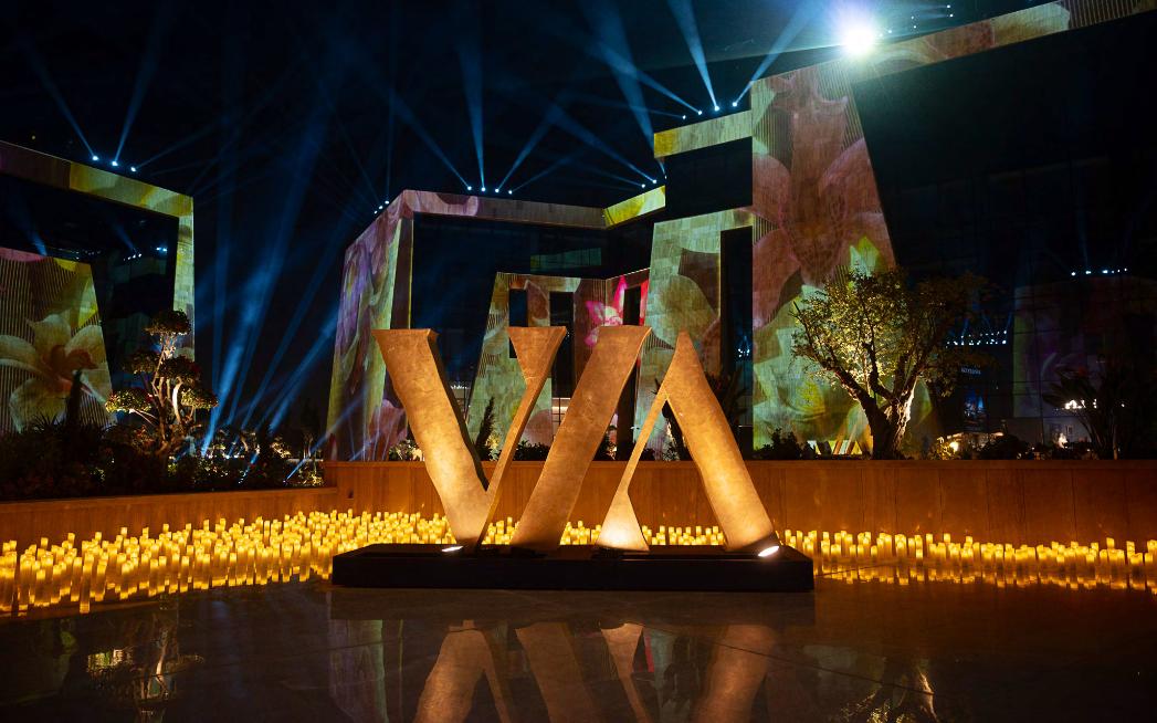 Monumental gold VIA letter sculpture ringed by hundreds of candles, with projection-mapped facades and skybeams behind, at the VIA Riyadh grand-opening gala.